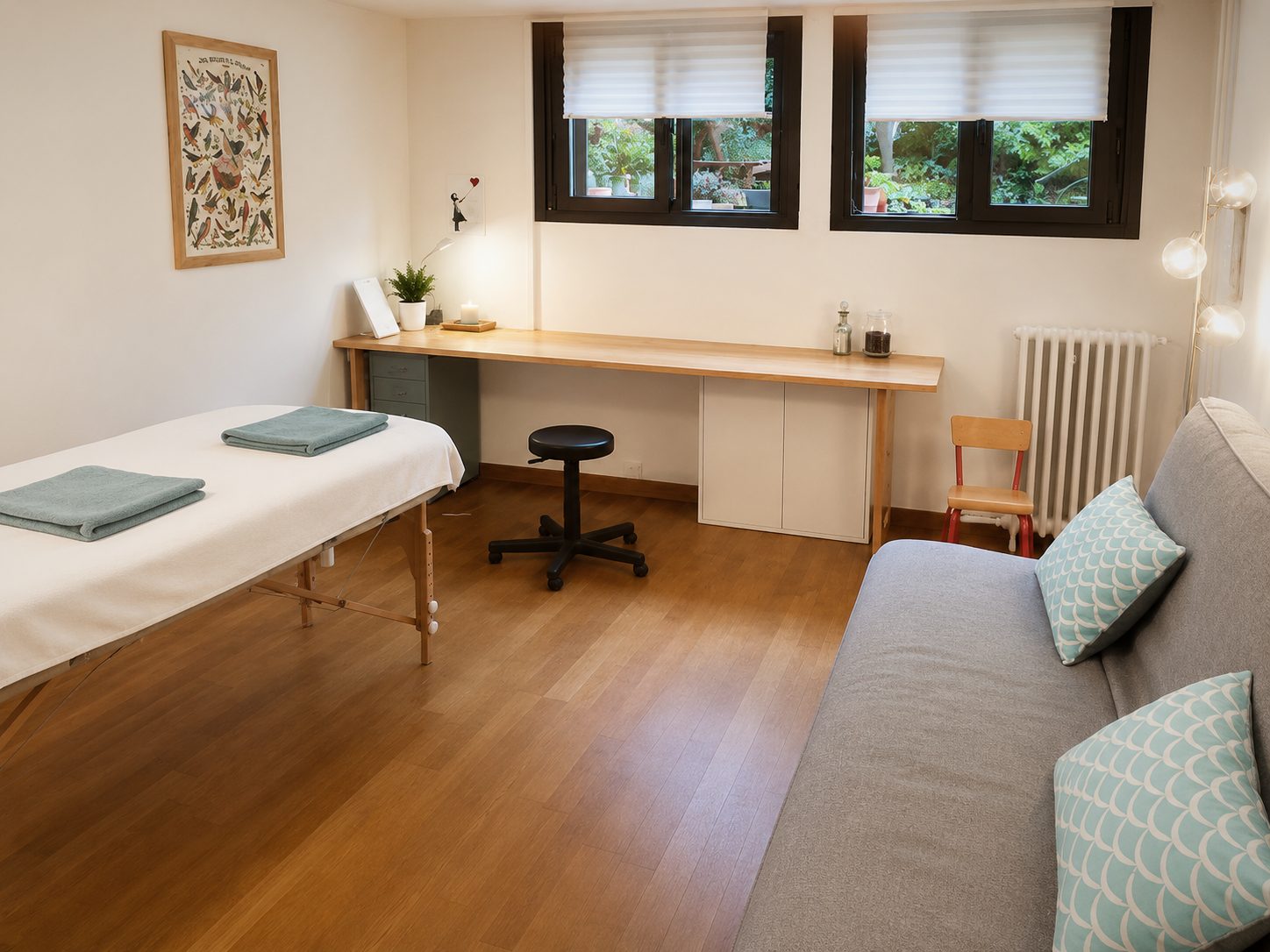 La pièce vue d'ensemble : la table de soin à gauche, un long plan de travail en bois sous deux fenêtres donnant sur un jardin, un canapé à droite.